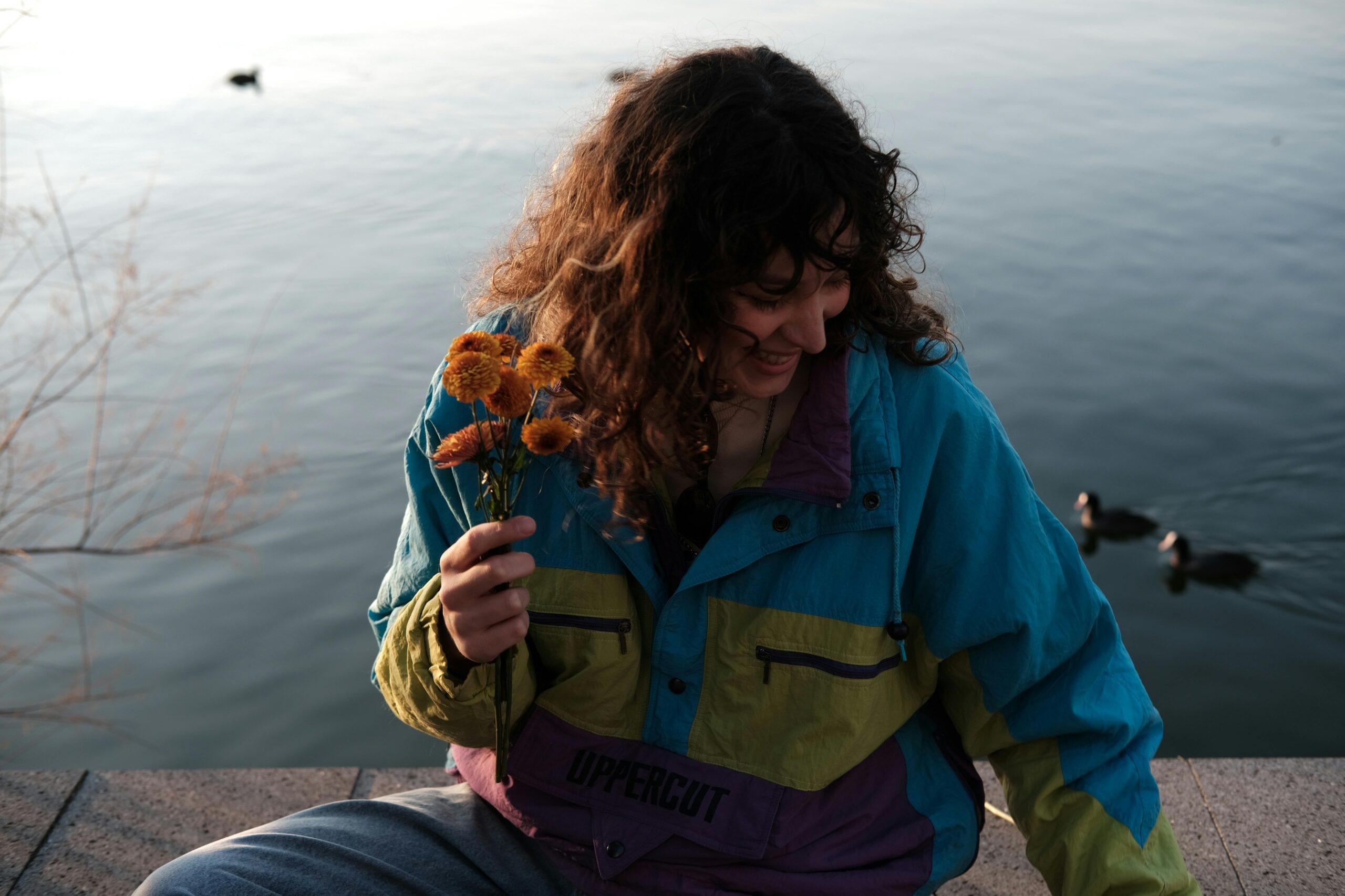 A joyful woman in a colorful jacket holding flowers by a serene lake with ducks.